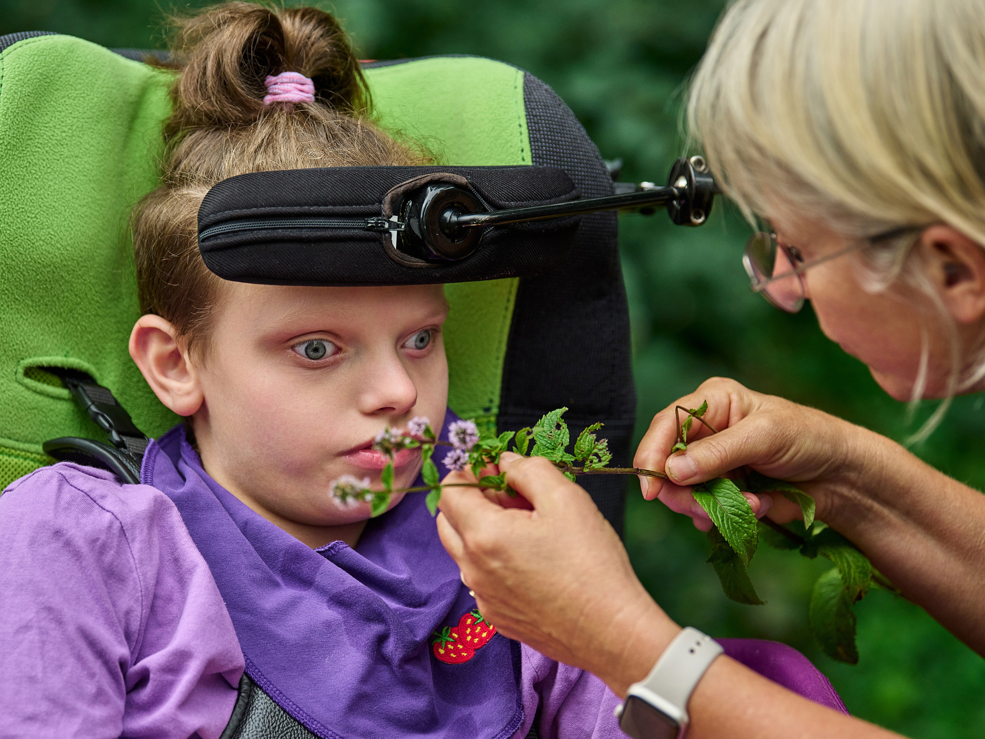 Teilhabe ermöglichen, Natur erfahren. Der sommerliches Kunstworkshop war für alle Beteiligten ein Erlebnis. Eine Betreuerin gibt einem Kind Kräuter zum Riechen.