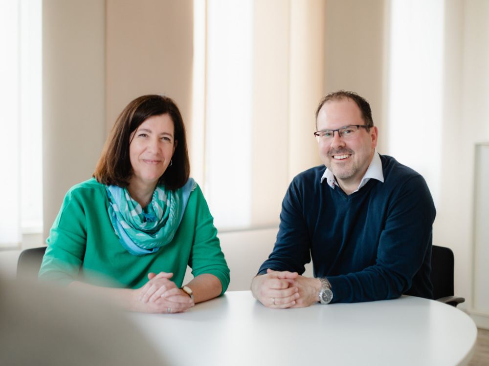 Mann und Frau sitzen lächelnd nebeneinander an einem Tisch.