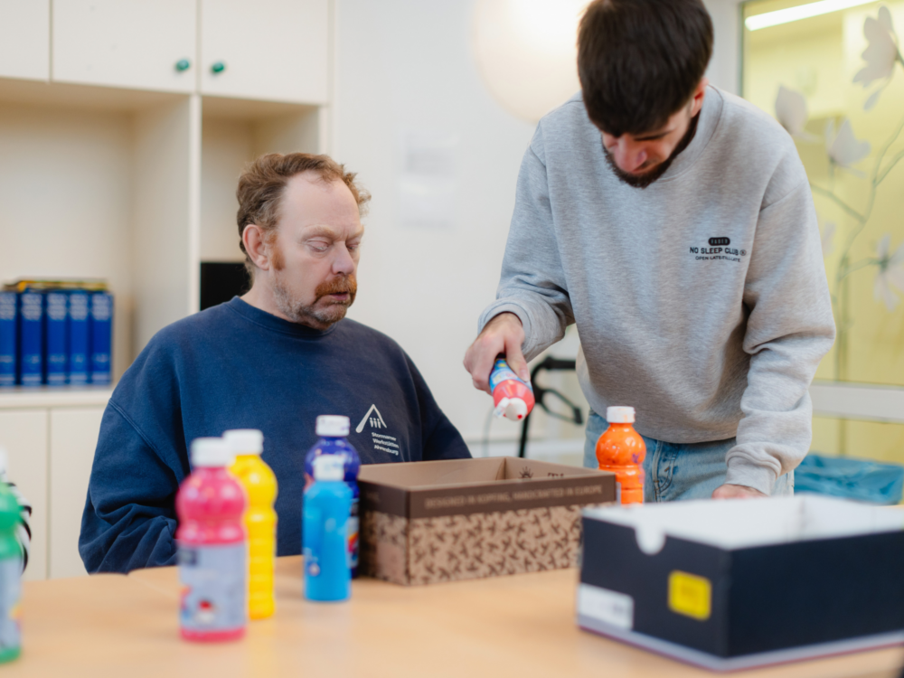 Ein junger Heilerziehungs-Fachschüler unterstützt einen Mann beim Malen.