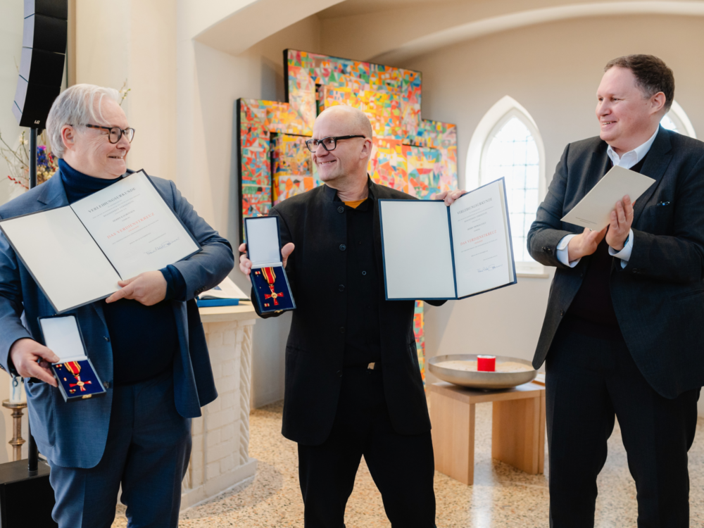 Kai Boysen und Thomas Cold bekommen von Kultursenator Dr. Carsten Brosda das Bundesverdienstkreuz überreicht. Die beiden lächeln sich gegenseitig an.