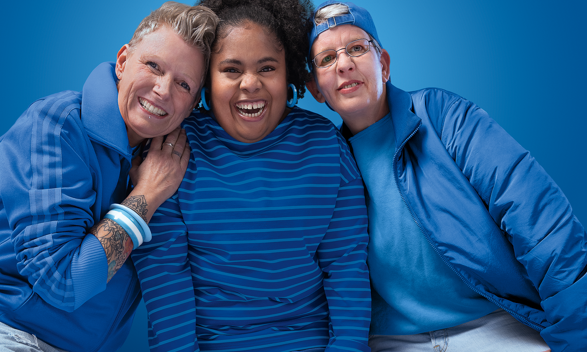 Kampagnen-Motiv: Drei Frauen lachen in die Kamera. Sie tragen blaue Kleidung und stehen vor einem blauen Hintergrund
