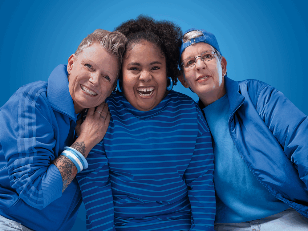 Kampagnen-Motiv: Drei Frauen lachen in die Kamera. Sie tragen blaue Kleidung und stehen vor einem blauen Hintergrund