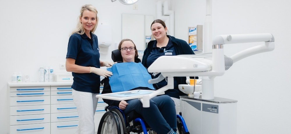 Zwei Personen stehen neben einem Zahnarztstuhl auf dem eine dritte Person sitzt