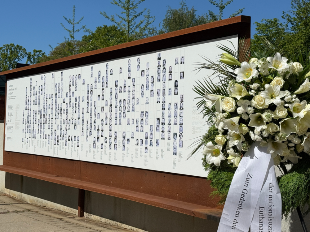 Ein Kranz aus Blumen liegt vor einer Tafel, wo Bilder und Namen von den Menschen stehen, die aus den Alsterdorfer Anstalten im zweiten Weltkrieg deportiert wurden.