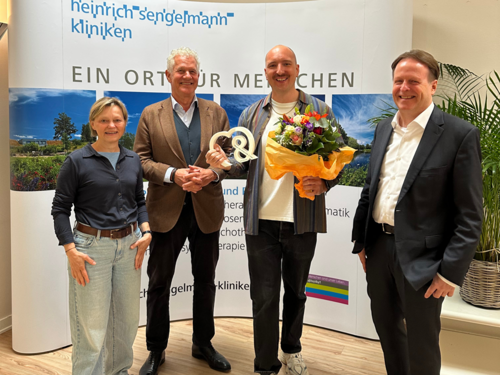 Vier Menschen lächeln in die Kamera. Ein Mann in der Mitte hält einen Preis und einen Blumenstrauß.