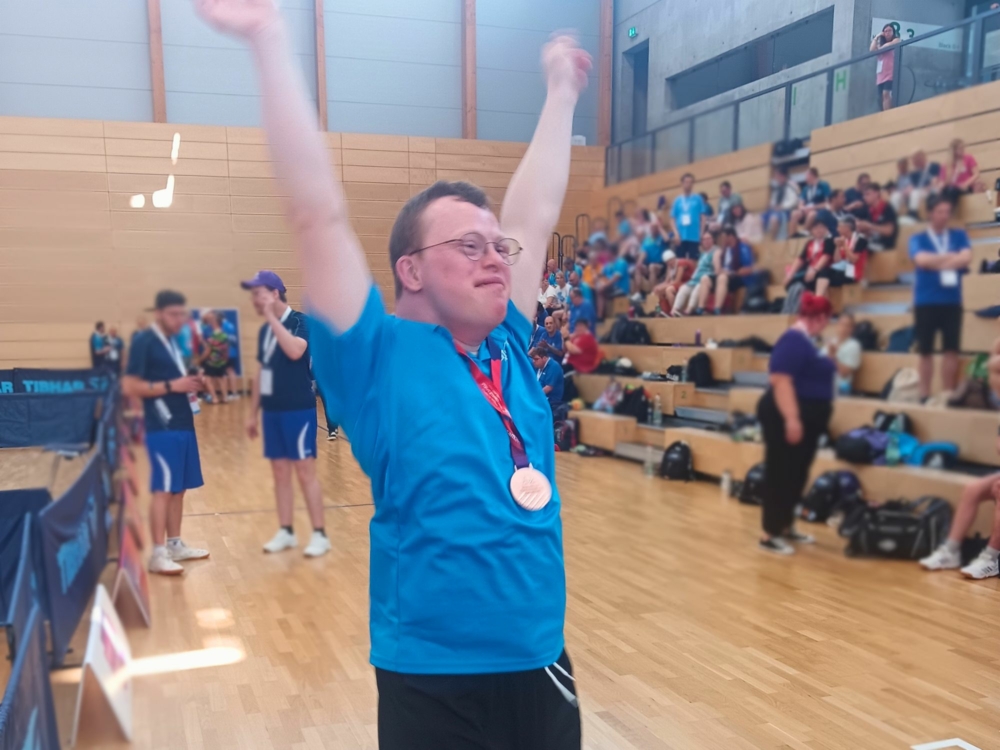 Ein Mann steckt die Arme in die Luft und freut sich über seine Bronze-Medaille. Er trägt eine Brille, ein blaues Shirt und kurze Haare.