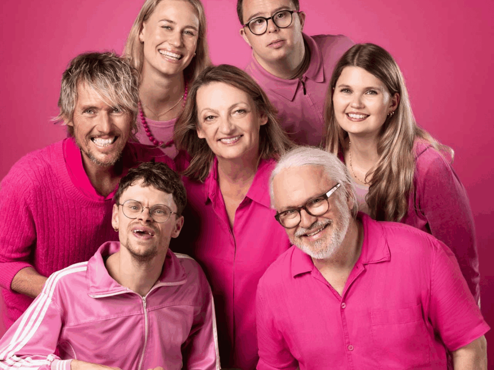 Sieben Menschen lachen in die Kamera. Einer sitzt auf seinem Rollator. Das ganze Bild ist von der Kleidung und dem Hintergrund her pink gehalten.
